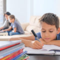 Junge macht lustlos Hausaufgabe, im Hintergrund ein Elternteil mit einem Kind am Laptop
