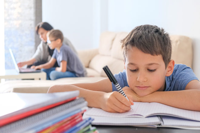 Junge macht lustlos Hausaufgabe, im Hintergrund ein Elternteil mit einem Kind am Laptop