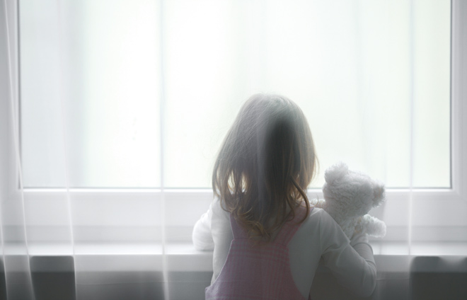Kind mit Teddy steht hinter einem Vorhang am Fenster