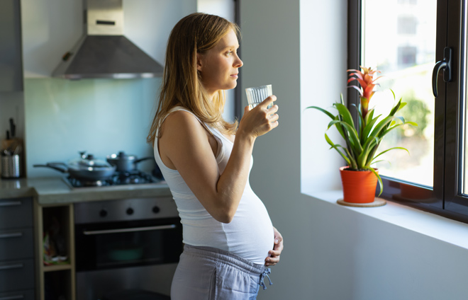 Schwangere Frau steht am Küchenfenster, hält eine Tasse in der Hand